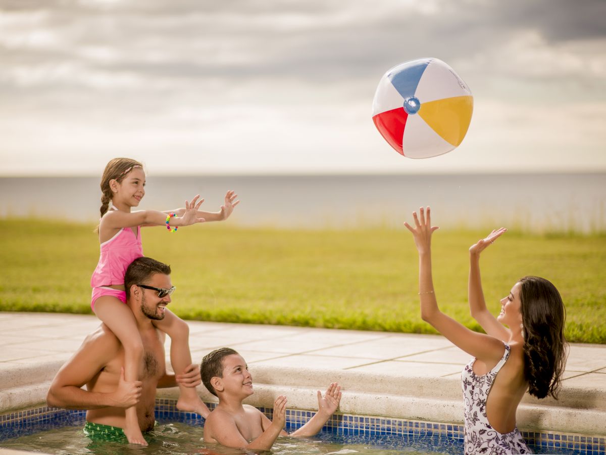 A family is enjoying time in a pool, playing with a beach ball. The father holds a child on his shoulders while the mother and another child play.