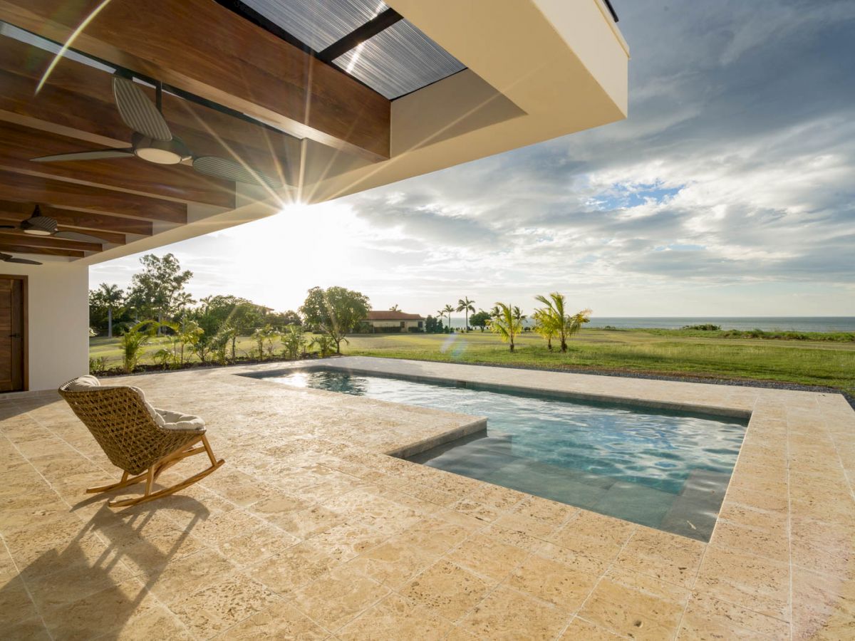 The image shows a luxurious outdoor space with a small pool, a wicker chair on a tiled patio, overlooking a grassy landscape and a view of the sea.