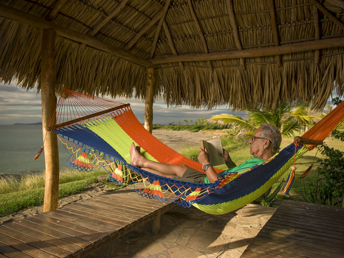 A man relaxes in a colorful hammock under a thatched roof, reading a book with a serene ocean view in the background.
