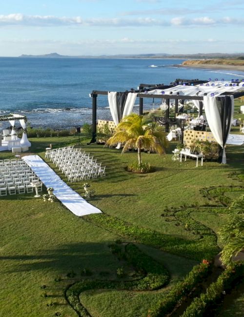 An outdoor beachside wedding setup with white chairs, an aisle runner, and decorated gazebos on a lawn, overlooking the ocean on a sunny day.
