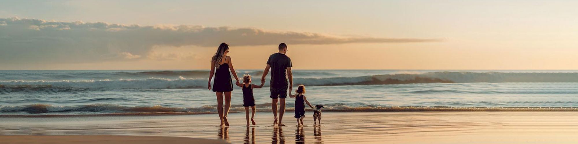 A family of four is walking hand-in-hand on a beach, heading toward the sea during sunset with their reflections in the wet sand.
