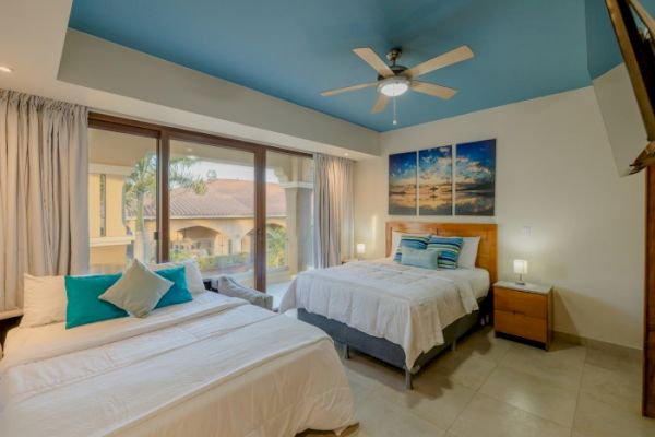 A modern bedroom featuring two beds with white linens, blue accent pillows, a ceiling fan, artwork on the wall, and a large window with a view.