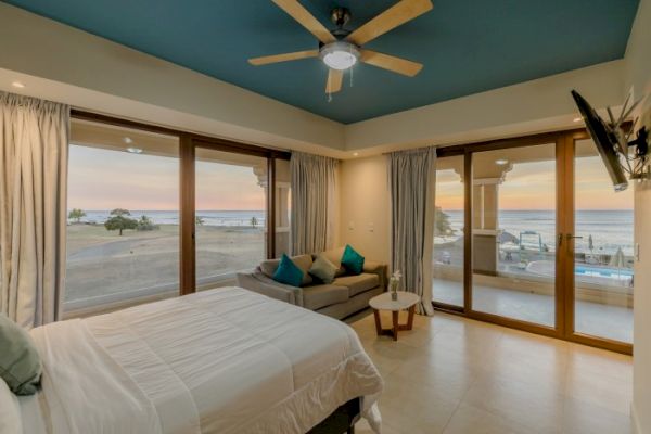 A modern bedroom with large windows offers a view of the sunset over the ocean, a bed, a sofa, a ceiling fan, and a mounted TV.