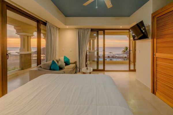 A cozy bedroom with a modern design featuring a bed, a small seating area, a wall-mounted TV, and large windows offering an ocean view at sunset.