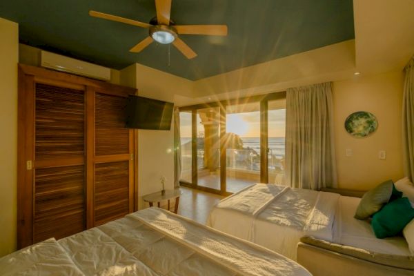 This image shows a sunlit bedroom with two beds, a ceiling fan, a wall-mounted TV, and large glass doors leading to a balcony overlooking a scenic view.