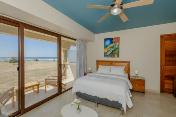 A bright bedroom with ocean view, features a double bed, wooden furniture, ceiling fan, and wall art, opening to a balcony with seating.