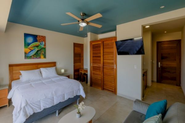 A modern bedroom with a bed, ceiling fan, wall-mounted TV, wooden closet, sofa, and vibrant artwork on the wall, featuring a blue ceiling.