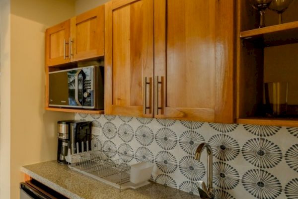 A kitchen with wooden cabinets, a microwave, coffee maker, dish rack, and a patterned tiled backsplash, ending the sentence.