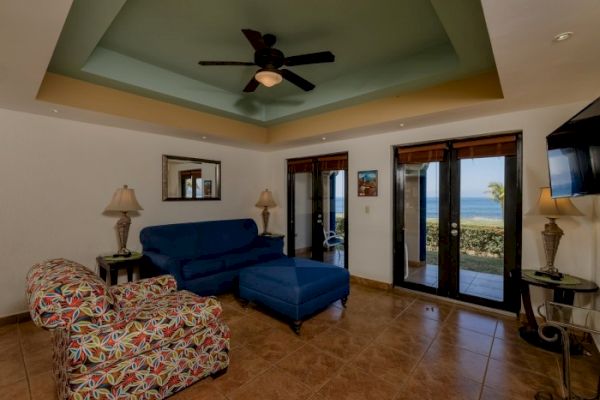 A cozy living room with colorful furniture, ceiling fan, and ocean view visible through glass doors, with tiled floors and wall decorations.