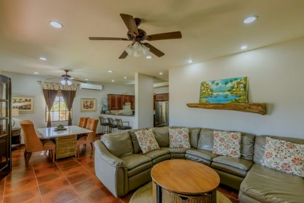 A cozy living room with a sectional sofa, a round coffee table, dining area, kitchen, ceiling fans, and wall art, all under warm recessed lighting.