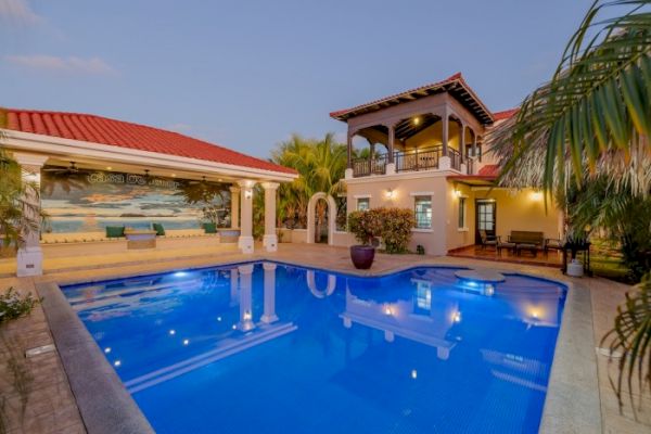 The image shows a luxurious house with a blue swimming pool, surrounded by palm trees and tropical landscaping, featuring a covered outdoor area.