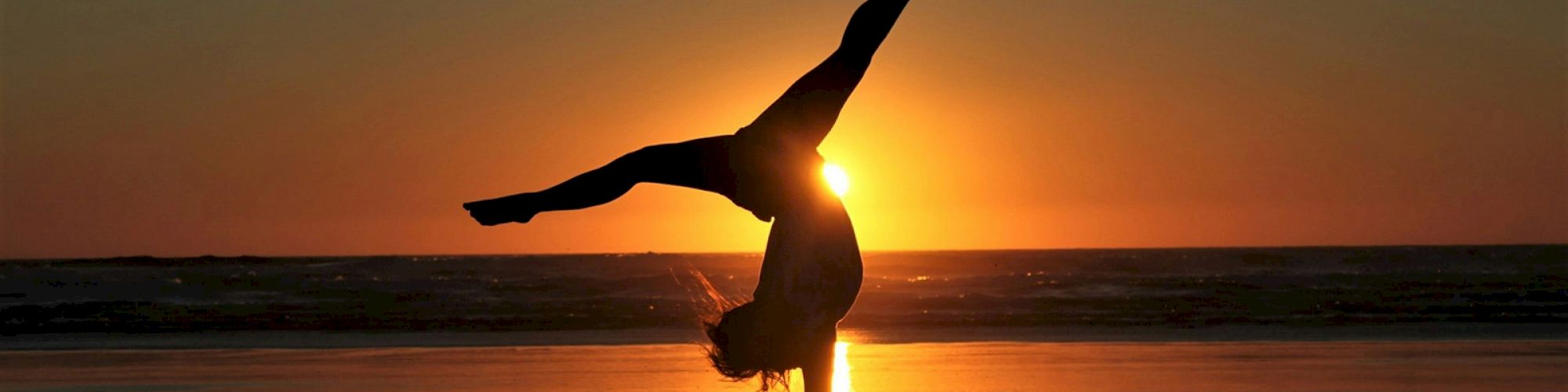 A person is doing a handstand on the beach during sunset, creating a striking silhouette against the vibrant sky and reflecting in the water.