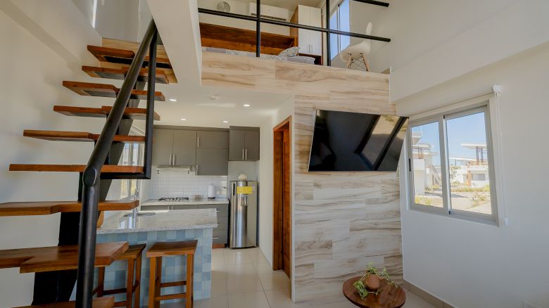 A modern loft apartment with a wooden staircase, kitchen, TV, and a small table with fruit. Bright natural light pours through the window.