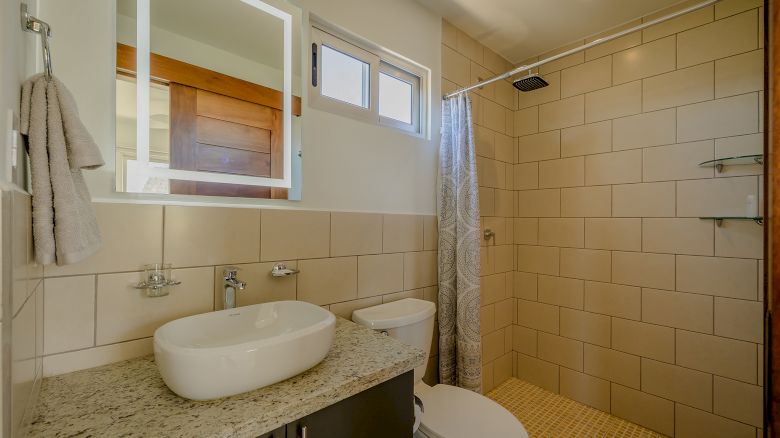 The image shows a modern bathroom with a granite countertop, a vessel sink, a toilet, a walk-in shower with a curtain, and beige tiles.