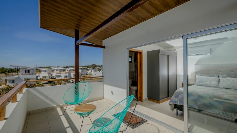 A modern balcony with two turquoise chairs and a small table, adjacent to a bedroom with a bed, and large glass sliding doors.