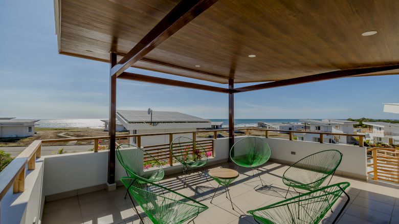 An outdoor patio with green wire chairs and a table, overlooking rooftops and the ocean, covered by a wooden ceiling.