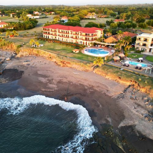 A coastal resort with a pool, several buildings surrounded by greenery, and a beach with waves breaking on the shore can be seen in the image.