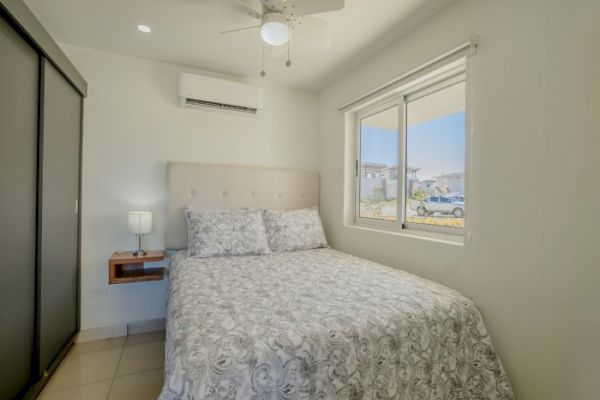 A small bedroom with a bed, nightstand, lamp, window, air conditioner, ceiling fan, and sliding door closet. The bed has a patterned comforter.