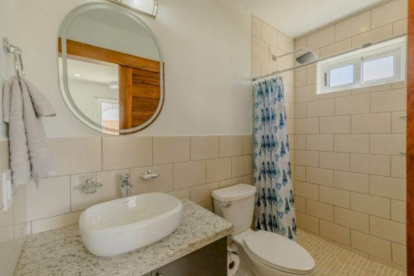 The image shows a bathroom with a round mirror, white sink on a granite countertop, a toilet, and a shower area with a blue patterned curtain.