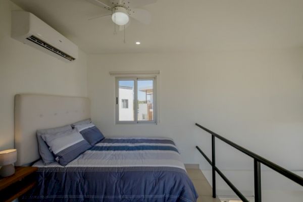 The image shows a modern bedroom with a bed, blue bedding, a headboard, a window, an air conditioner, and a ceiling fan, adjacent to a staircase.