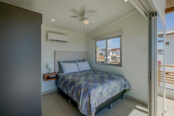 The image shows a modern bedroom with a queen-size bed, a window, air conditioning, a ceiling fan, and a sliding door leading to a balcony area.