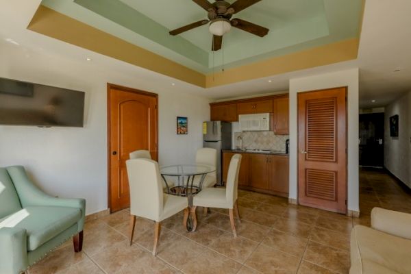 The image shows a cozy room with a ceiling fan, a dining table with chairs, a flat-screen TV, armchairs, and a small kitchen area with wooden cabinets.