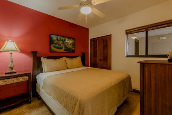 A cozy bedroom with a large bed, red accent wall, bedside lamp, painting, ceiling fan, dresser, and closet with louvered doors.