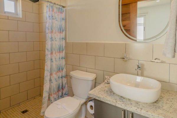 A bathroom with a shower, toilet, round mirror, floral shower curtain, countertop sink, and a small window.