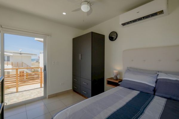 A modern bedroom with a bed, wardrobe, nightstand, ceiling fan, and air conditioner, leading to a balcony through a sliding glass door.