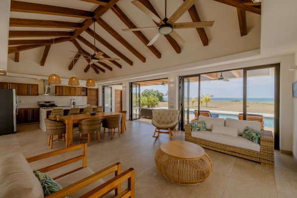 A spacious living area with wooden beams, ceiling fans, and modern furniture, featuring an open kitchen and large windows offering a view of the ocean.