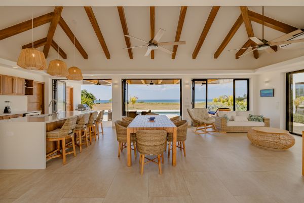 A spacious open-plan living area with a wooden-beamed ceiling, wicker furniture, and a view of the ocean through large sliding glass doors.