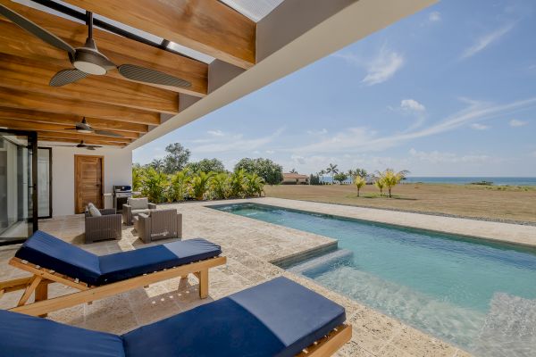 A luxurious poolside area with lounge chairs, a wooden ceiling with fans, an outdoor seating area, surrounded by lush greenery, and a distant ocean view.