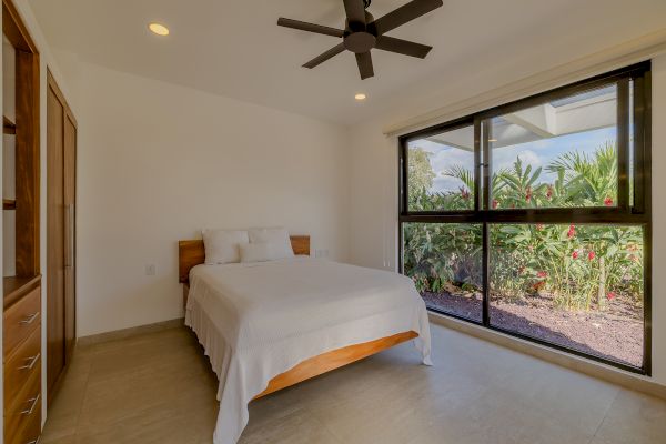 A minimalist bedroom with a large window, a ceiling fan, a bed with white linens, and a view of lush greenery outside the window.