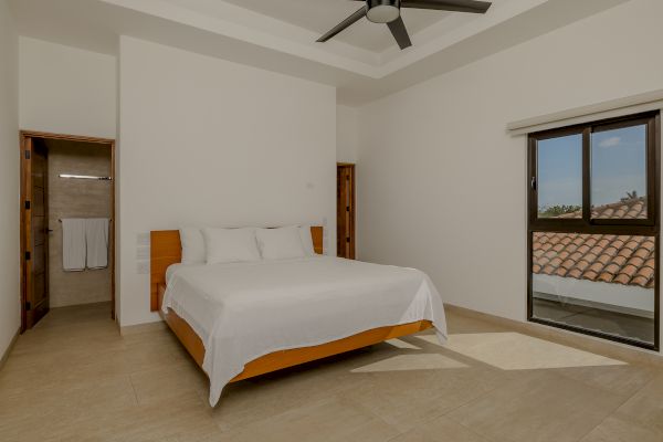 A bedroom features a bed with white linens, a ceiling fan, a window with a view, and an open doorway leading to a bathroom with towels hung.