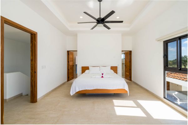 A minimalist bedroom with a bed, white linens, orange accents, ceiling fan, two doors on either side, and a window with a view outside.