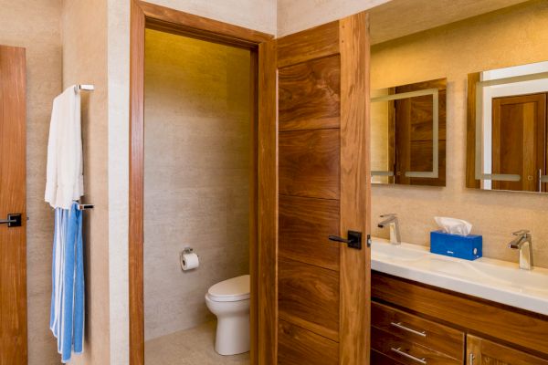 A bathroom with a wooden door leading to a toilet, a sink with a large mirror, wooden cabinets, and towels hanging on the wall.