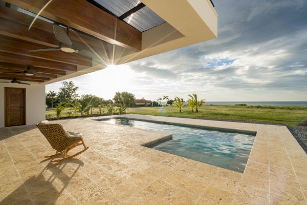 A luxurious outdoor pool area with a wicker chair, overlooking an expansive green landscape and water under a partly cloudy sky and sunshine.