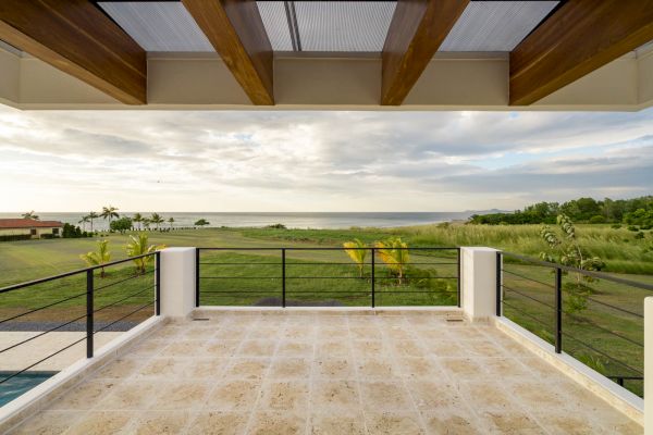 A spacious terrace overlooking a grassy field with palm trees, and a view of the ocean under a partly cloudy sky, framed with wooden beams.