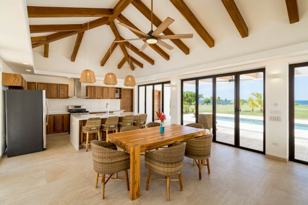 This image shows a modern open kitchen and dining area with wooden beams, wicker chairs, a wooden table, and large windows offering a view of a pool and garden.