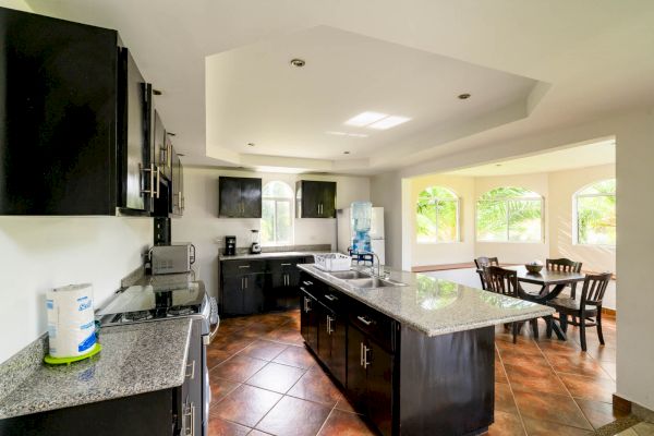 A modern kitchen with dark cabinets, granite countertops, stainless steel appliances, and an adjacent dining area with large windows and a water dispenser.