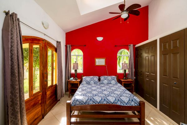 The image shows a bedroom with a red accent wall, a bed in the center, two nightstands, a ceiling fan, large wooden doors, and closets.