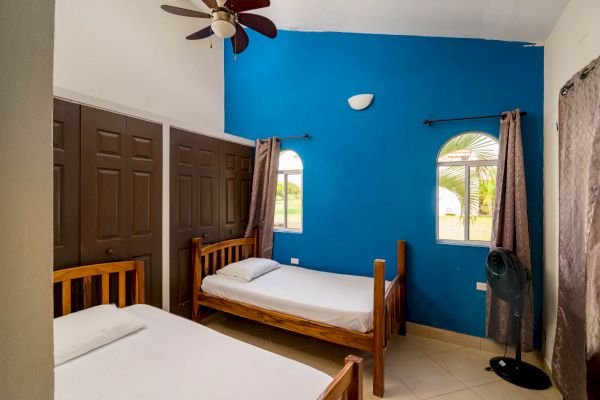 A room with two single beds, brown closets, a ceiling fan, blue accent wall, two windows with curtains, and a standing fan.