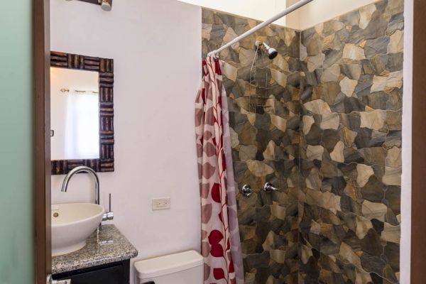 This image shows a small bathroom with a sink, mirror, toilet, and a stone-tiled shower area with a curtain.