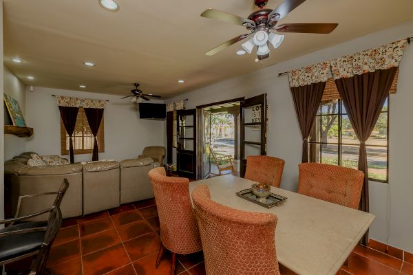 A cozy living room and dining area with a sofa, dining table, ceiling fans, and large windows with curtains, featuring a warm and inviting atmosphere.