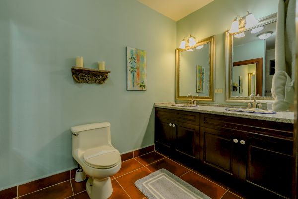 The image shows a bathroom with a toilet, double sink vanity, large mirror, wall décor with candles, artwork, and two wall-mounted lights.