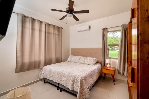 A cozy bedroom with a double bed, beige curtains, a ceiling fan, an air conditioner, and a bedside table with a lamp next to a window.
