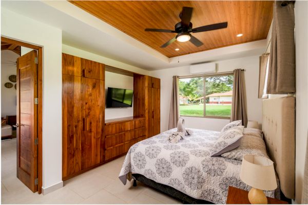 This image shows a cozy bedroom with a wooden ceiling, a ceiling fan, a large window, a bed with patterned bedding, and a wooden wardrobe with a TV.