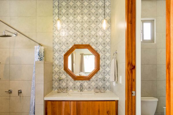 A bathroom with a wooden-framed mirror, pendant lights, a wooden vanity, a walk-in shower, a toilet, and patterned tiles near the sink area.