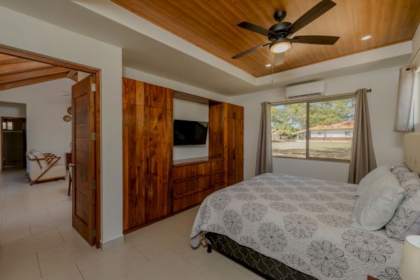 This image shows a cozy bedroom with a patterned gray bedspread, a wooden wardrobe with a TV, a ceiling fan, and a window with a view.