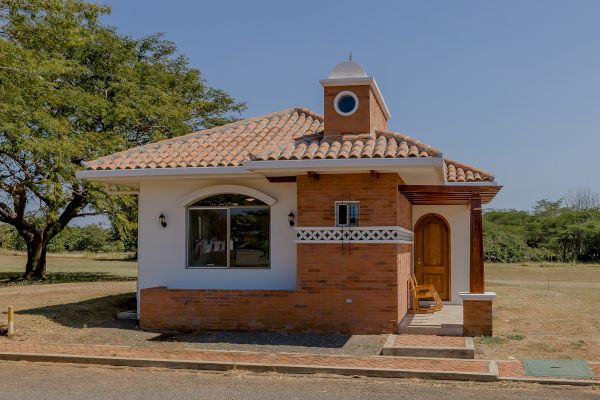 A small, charming brick and stucco house with a tiled roof and arched windows, situated in a grassy area with trees.